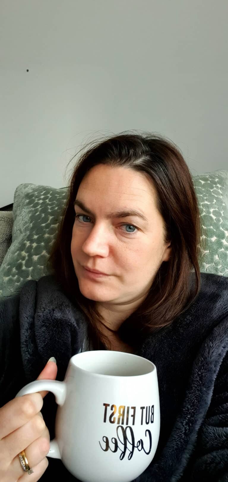 A woman with brown hair sits on a patterned cushion, wearing a dark dressing gown and holding a white mug that says BUT FIRST Coffee. She looks at the camera with a neutral expression, perhaps reflecting on reasons for emotional reactions.