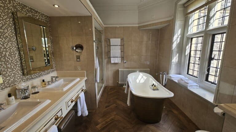A luxurious large bathroom features elegant bathroom decor, including a freestanding bath, double basins with a large mirror, a glass-enclosed shower, and large windows. Towels are neatly arranged on racks and shelves for stylish bathroom styling.