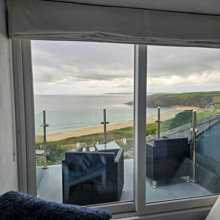 View through a glass door with modern cladding, showing a balcony with two chairs overlooking a sandy beach, calm sea, and green hills under a cloudy sky.