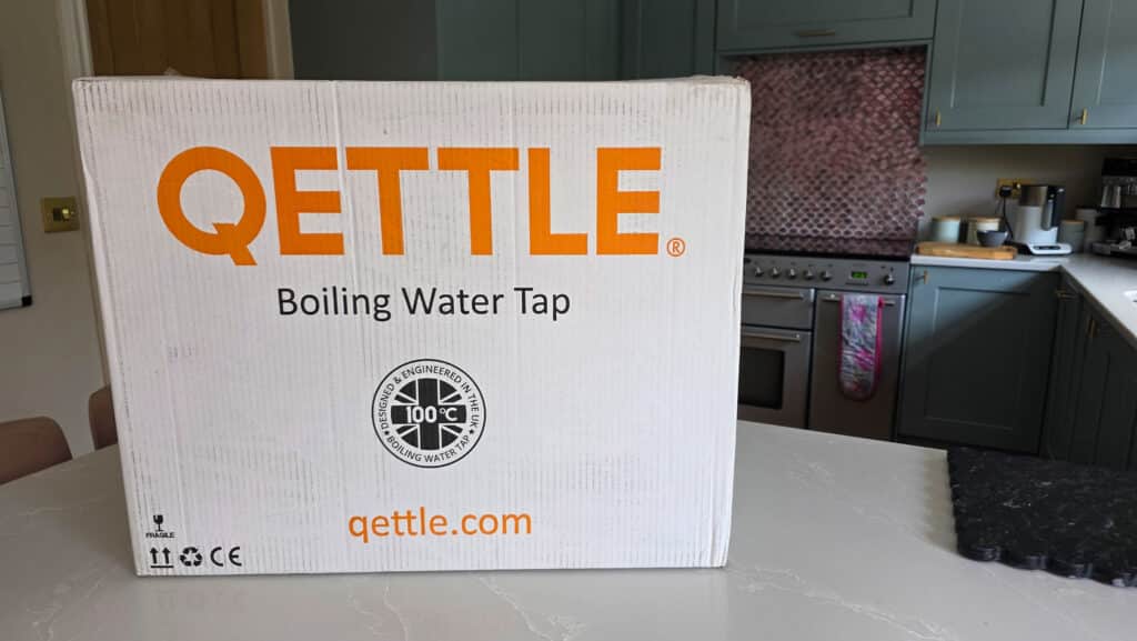 A large white box labelled QETTLE Boiling Water Tap sits on a kitchen worktop, promising instant hot water. Kitchen cupboards, an oven, and appliances are visible in the background.