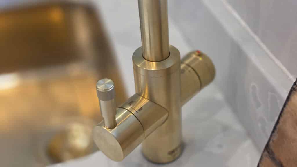 Close-up of a modern brushed gold kitchen tap with a cylindrical handle and temperature control, featuring instant hot water functionality. The background shows part of a gold sink and white worktop.