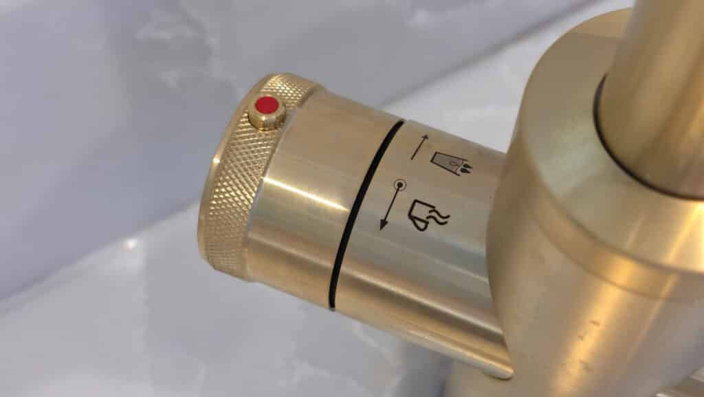 A close-up of a metallic, gold-coloured QETTLE boiling water tap handle with a red button and symbols for hot and cold water. The textured grip is visible, and the blurred background adds focus—perfect for those seeking instant hot water.