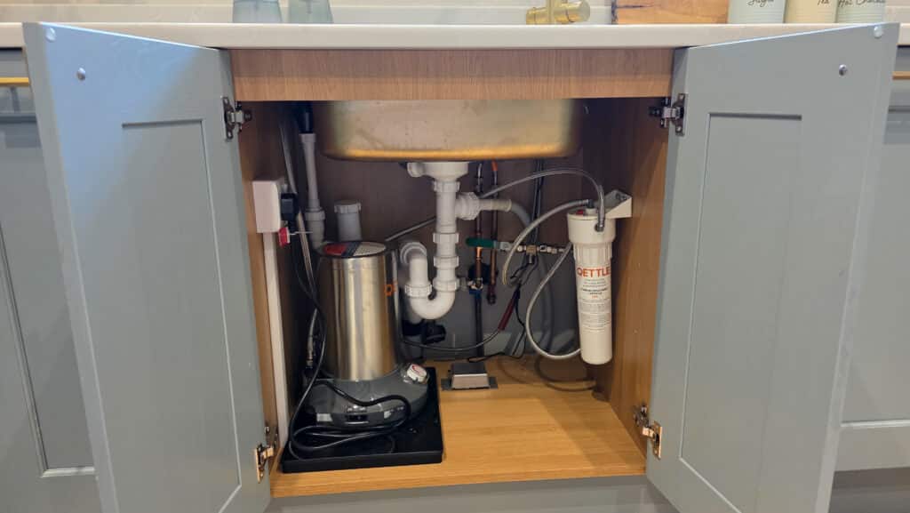 Open cupboard showing the underside of a kitchen sink with exposed plumbing pipes, a water filtration system, and a QETTLE Boiling Water Tap among various fittings and electronic components installed inside the cupboard.