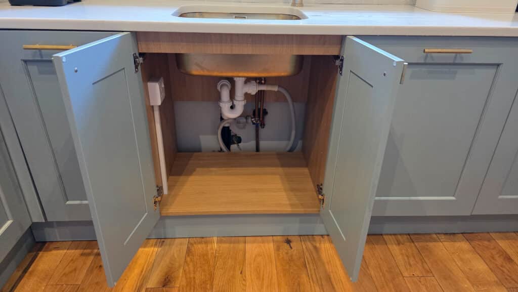Open kitchen cupboard under a sink showing exposed plumbing pipes, valves, and a QETTLE boiling water tap inside, with wooden interior and light blue cupboard doors on a wooden floor.