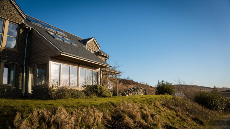 A modern house with large windows and a sloped roof sits on a grassy hill under a clear blue sky, featuring rooflights that add light to its compact rooms amid shrubs and distant trees.