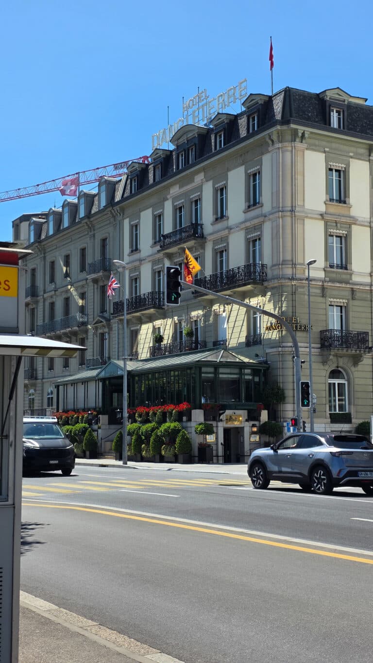 A large historic hotel with cream-coloured walls, black railings, and flags on the façade stands near a busy street—reminding us that buyers’ mistakes can happen if you overlook location when buying property abroad. Flowers decorate the balcony and entrance.