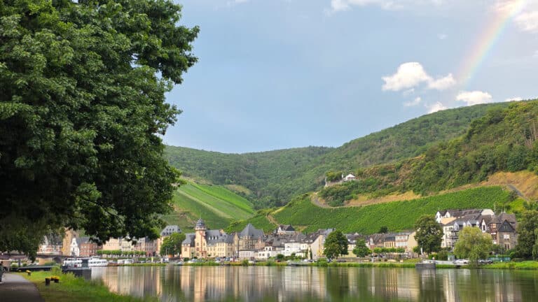 A quaint village with pastel-coloured buildings sits beside a calm river, surrounded by lush green hills and vineyards—one of those unexpected destinations where every view feels magical. A tree frames the scene, with a faint rainbow in the blue sky above.
