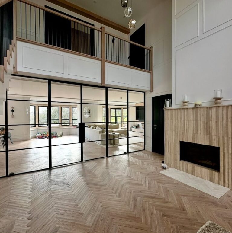 A spacious modern living area with herringbone wood floors, a glass wall with black frames, internal steel doors, high ceilings, a beige fireplace, and an open upstairs balcony overlooking a bright, furnished room.