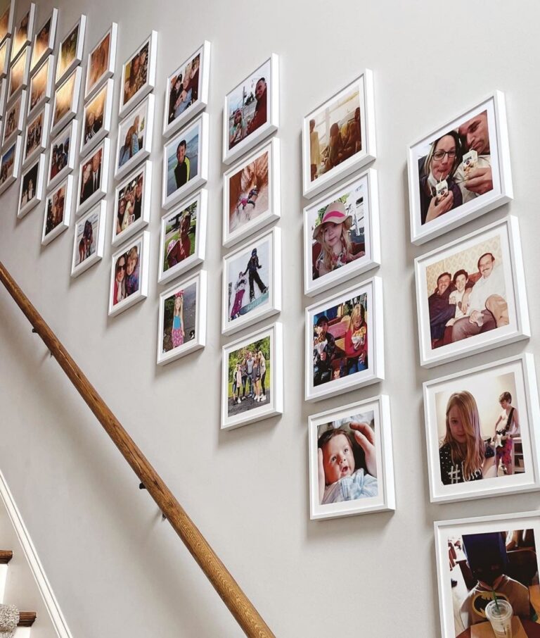 A collection of framed family photos arranged in a grid along a staircase wall, showing people of various ages in candid and posed moments. The white frames evoke the charm of a family-friendly vegetable garden, evenly spaced above the wooden bannister.