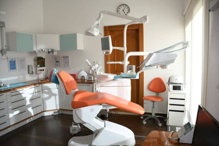A modern, sunlit dental surgery with an orange dental chair, overhead light, and various dental equipment showcases advanced modern dental technology. Cupboards and drawers line the walls, with a clock above a wooden door in the background.
