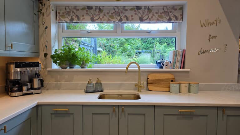 A bright kitchen with pale blue cupboards, a gold tap, a coffee machine, herb pots on the windowsill, and “What’s for dinner?” written on the wall. A large window overlooks a green garden.