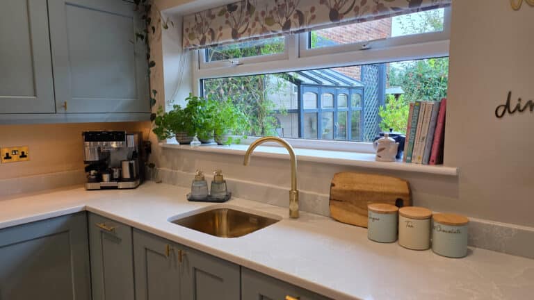 A modern kitchen with light blue cupboards, a brass tap, and a white worktop. Herbs, books, and decor rest on the windowsill, while jars labelled sugar, tea, and biscuits sit on the counter. A coffee machine is tucked in the corner.
