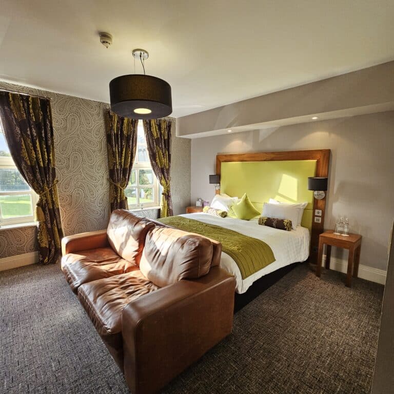 A sunlit hotel room features a large bed with green accents, a brown leather sofa at its foot, patterned wallpaper, and two windows—perfect for relaxing after pre-booking your airport taxi for a seamless travel experience.