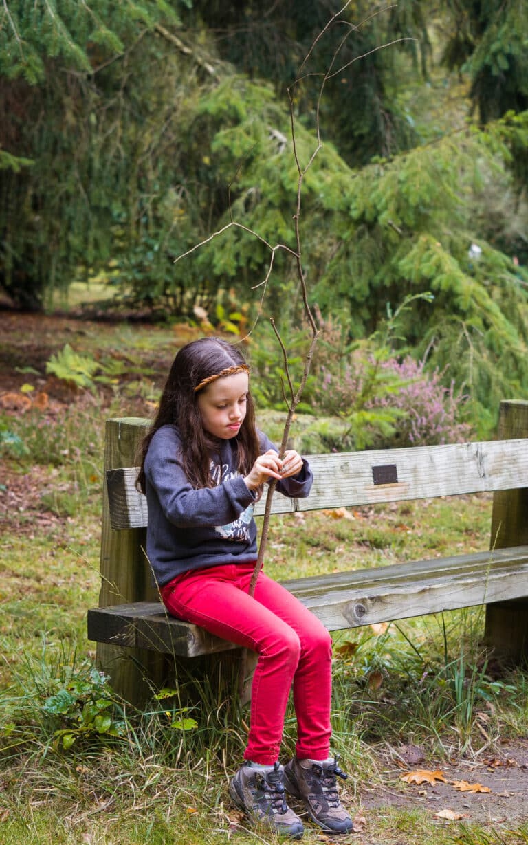 A young girl with long brown hair sits on a wooden bench in a park, her winter skin contrasting with a dark jumper and red trousers, as she holds and examines a long, thin tree branch amid lush green trees and grass.