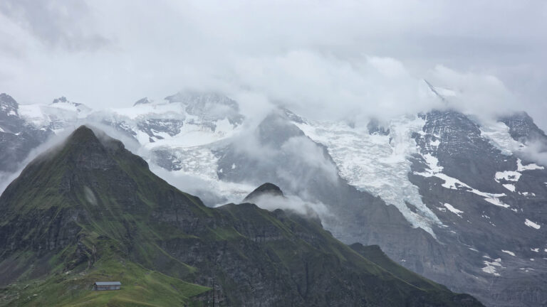 Make The Most of Your Short Winter Getaway Clouds and mist partially cover jagged, snow-capped mountains—an inviting scene for a Winter Getaway. In the foreground, a steep green hill slopes down to a small building, contrasting with rocky peaks and glaciers in the background.