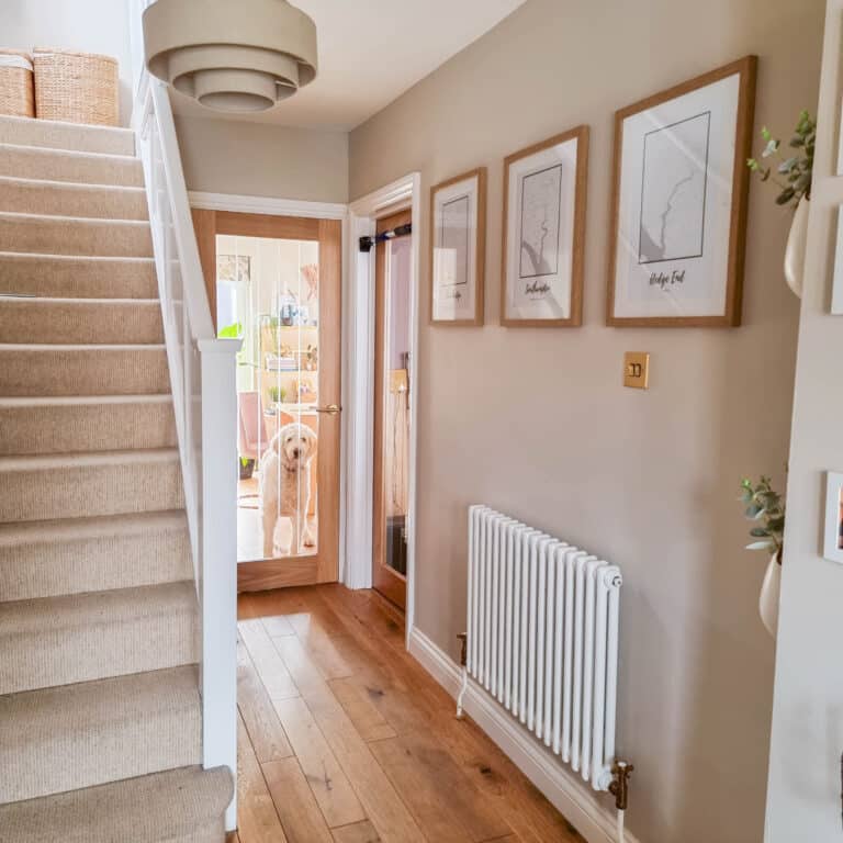 A bright hallway with wooden floors, beige walls, a staircase with carpet, bespoke wall art featuring framed maps, and a white dog standing behind a glass door at the end of the hallway.