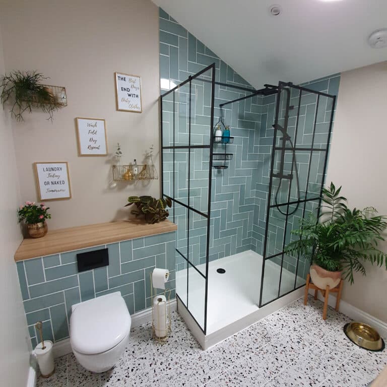This modern bathroom features a light blue tiled shower, black-framed glass doors, wooden shelf with decor and plants, white toilet, terrazzo floor, wall art, gold accessories, and lush greenery for a chic finishing touch.