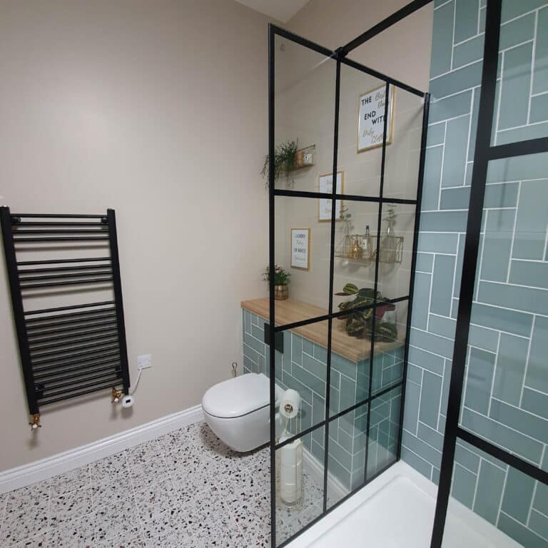 Modern bathroom with terrazzo flooring, green geometric tiles in the shower, a floating toilet, a sleek UK towel rail, and a wooden countertop with plants and decor behind a black-framed glass partition.