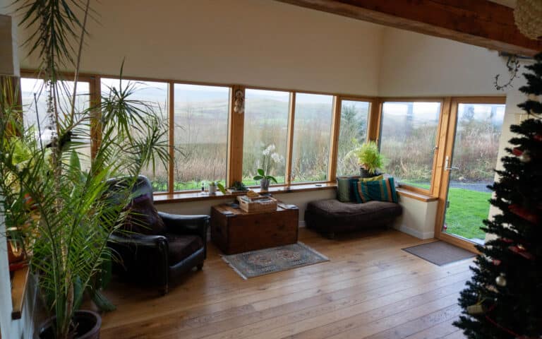 A bright sunroom with large windows overlooking a grassy landscape, wooden floors, potted plants, a leather armchair, cushioned bench, wooden chest, and Christmas tree—perfect for clients searching with Estate Agents in Peterborough.