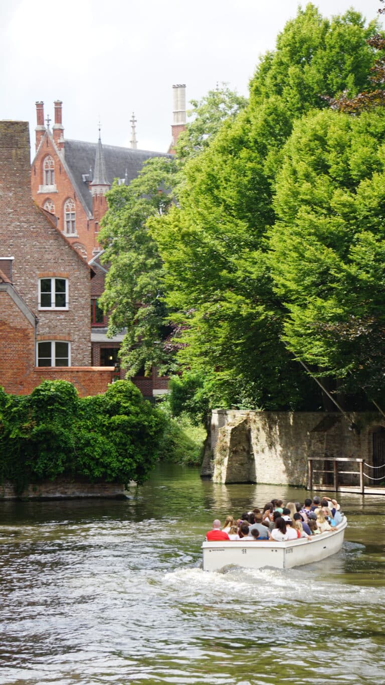 A small boat filled with people travels along a canal, bordered by lush green trees and historic brick buildings under a bright sky.