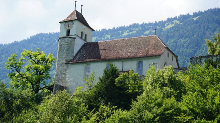 A stone church with a clock tower and red-tiled roof stands on a hill, surrounded by lush green trees—a picturesque scene reminiscent of prime property syndication opportunities, set against forested mountains under a bright, cloudy sky.