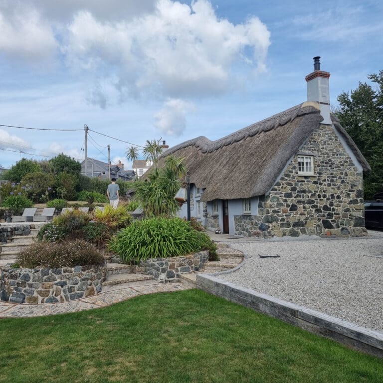 A charming stone cottage with a thatched roof and Best Timber Windows, surrounded by gardens and greenery under a partly cloudy sky. A person stands near the bushes in the landscaped garden.