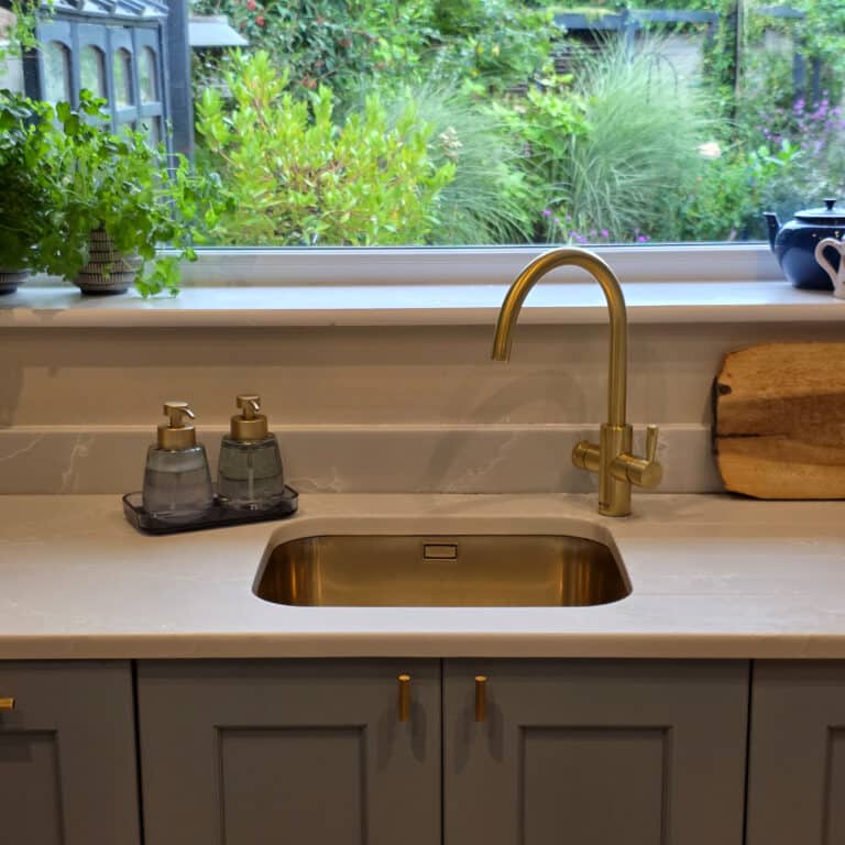 Modern kitchen sink with gold tap, soap dispensers, and wooden chopping board on a white worktop. Lush plants and a window with a garden view complete the look—ideal for preventing water damage left untreated in your home.
