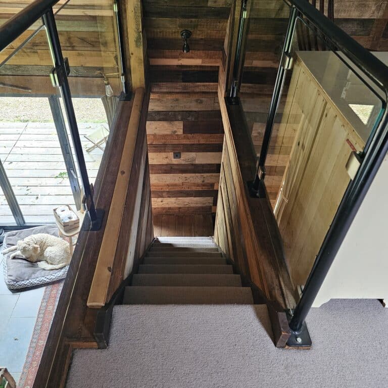 View looking down a wooden staircase with glass balustrades. At the bottom is a wooden feature wall. To the left, a dog lies curled up on a bed near a glass door leading to an outdoor decking.