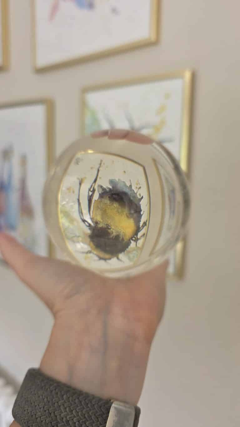 A hand holds a glass sphere with a black and yellow abstract shape inside, resembling a splash of paint—much like the mysterious visuals on the best online psychic platforms. Blurred framed art pieces hang in the background on a beige wall.