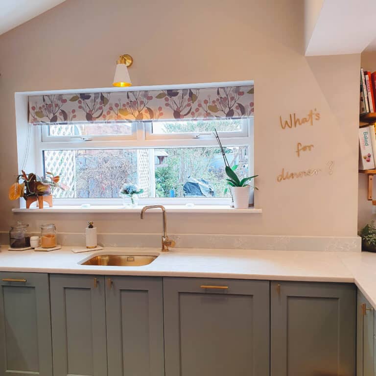 Modern kitchen with blue-grey lower cabinets, white worktop, and a sink beneath a window. The window has a floral-patterned blind and potted plants. What's for dinner? is written on the wall in gold letters.
