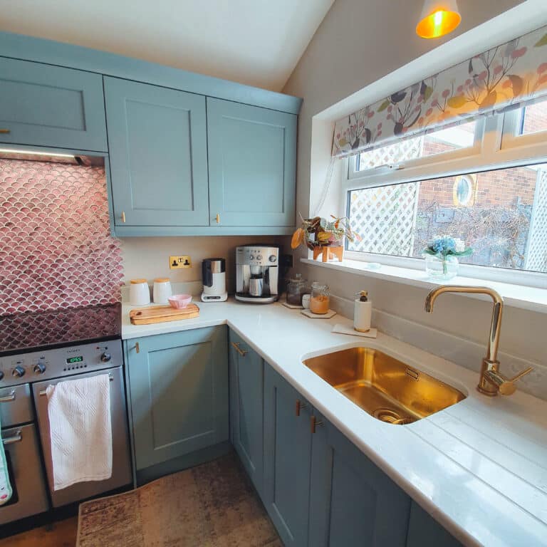 A cosy kitchen with light blue cupboards, a gold sink and tap, white worktops, a cooker, coffee maker, and a window with a floral Roman blind letting in natural light.