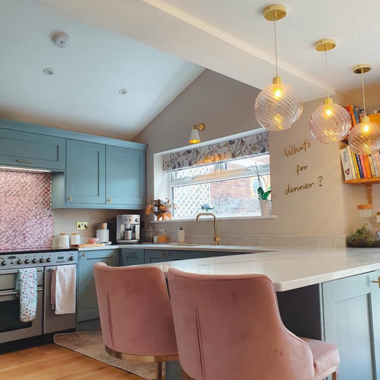 Modern kitchen with blue cupboards, a marble worktop island, two pink velvet chairs, three hanging globe lights, and a note on the wall reading “What’s for dinner?” A window lets in natural light above the sink.