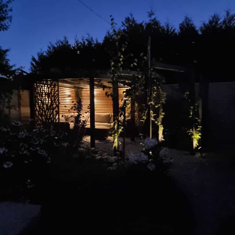 A cosy wooden gazebo lit with warm lights stands in a garden at dusk, surrounded by trees and plants with softly illuminated greenery—a perfect escape after tackling boiler problems that lead to replacement.
