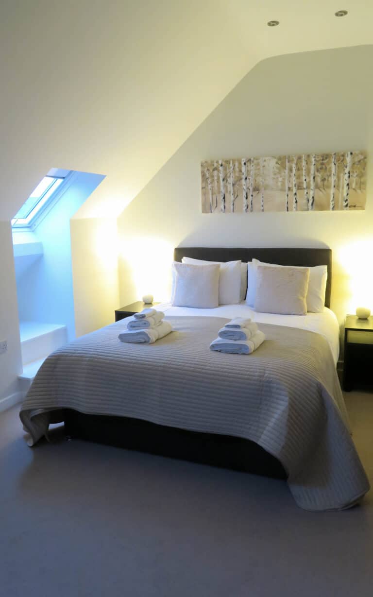A neatly made double bed with folded towels on top, flanked by two lit bedside lamps. The room, reflecting the advantages of buying a new-build, has cream walls, an angled ceiling with a skylight, and artwork above the headboard.