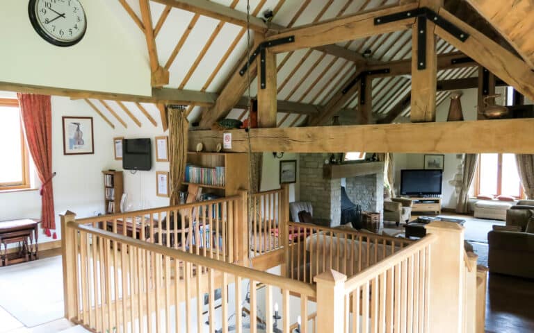 A spacious, rustic living room with exposed wooden beams, a stone fireplace, bookshelves, and armchairs features a safe staircase design with a sturdy wooden bannister. Large windows with red curtains fill the room with natural light.