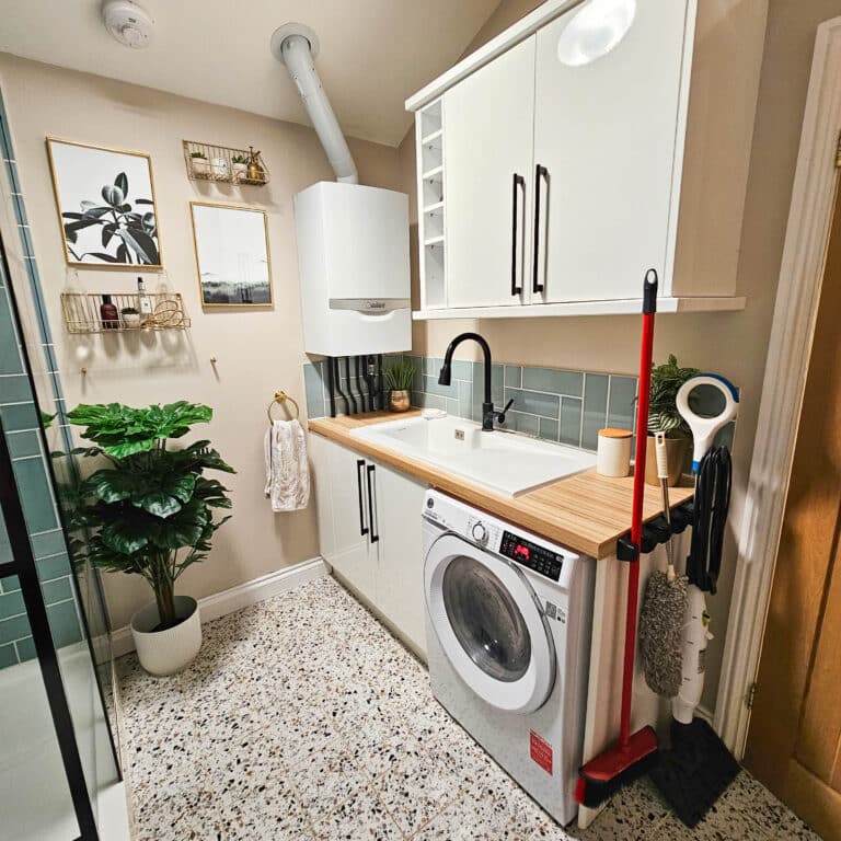 Modern laundry room with a washing machine under a wooden worktop, white cupboards above, green tiled splashback, potted plants, and wall art. Proper care prevents boiler problems that lead to replacement in spaces like this. Speckled flooring adds charm.