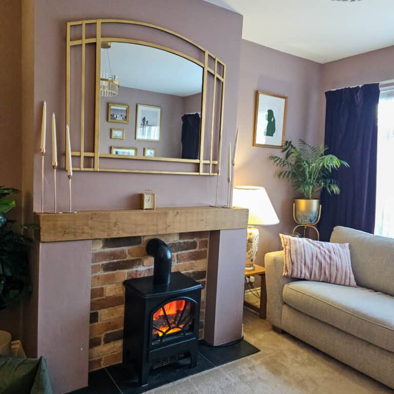 A cosy living room with a beige sofa, striped cushion, black wood stove, brick fireplace, tall gold candles, a large gold-framed mirror, framed art, a plant, and a lit table lamp on a wooden mantelpiece—ideal for relaxing after researching the Best Energy-Efficient Water Pumps.
