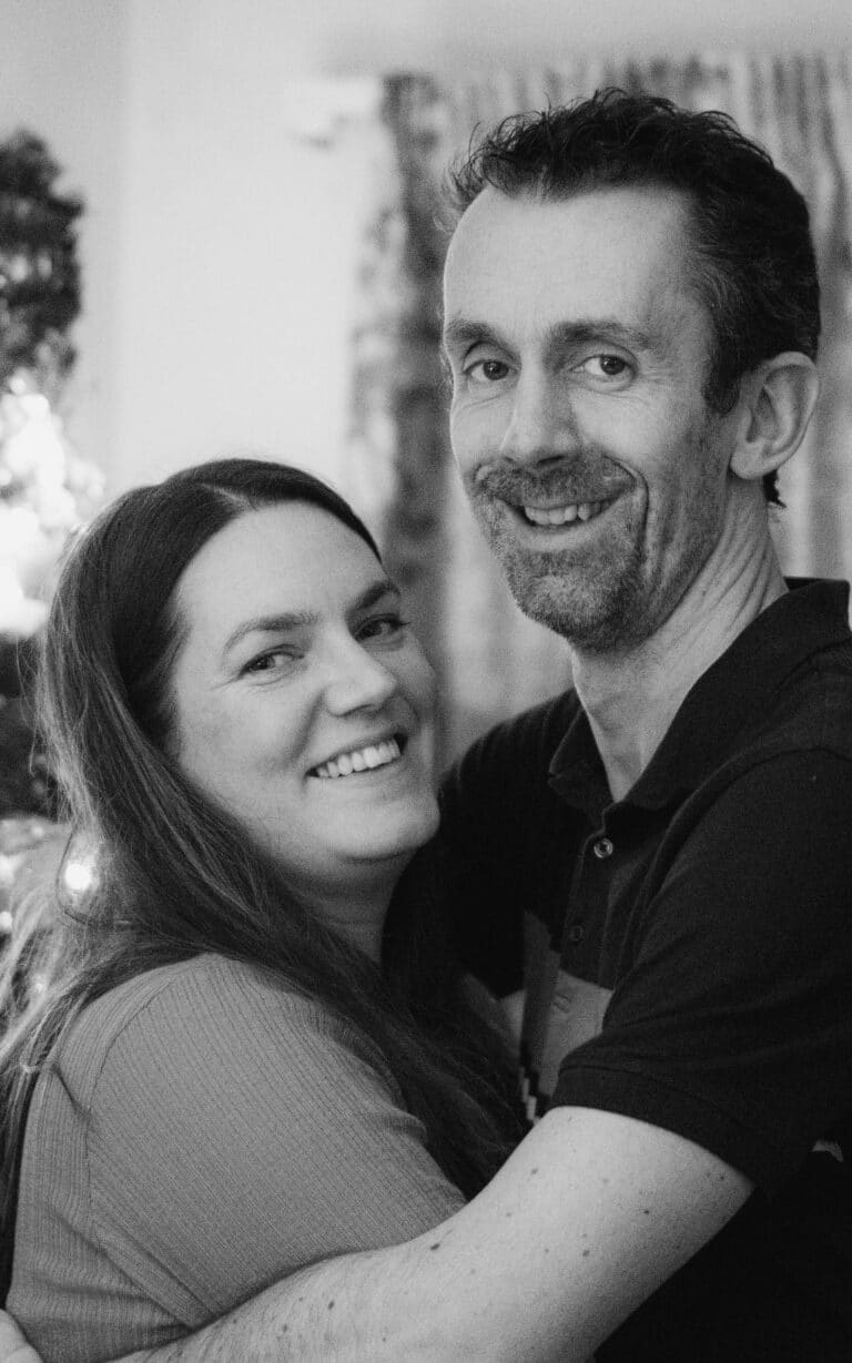 A black-and-white photo of a smiling woman and man embracing each other indoors, with soft lighting highlighting her hand that subtly showcases elegant engagement ring designs against the backdrop of soft curtains.