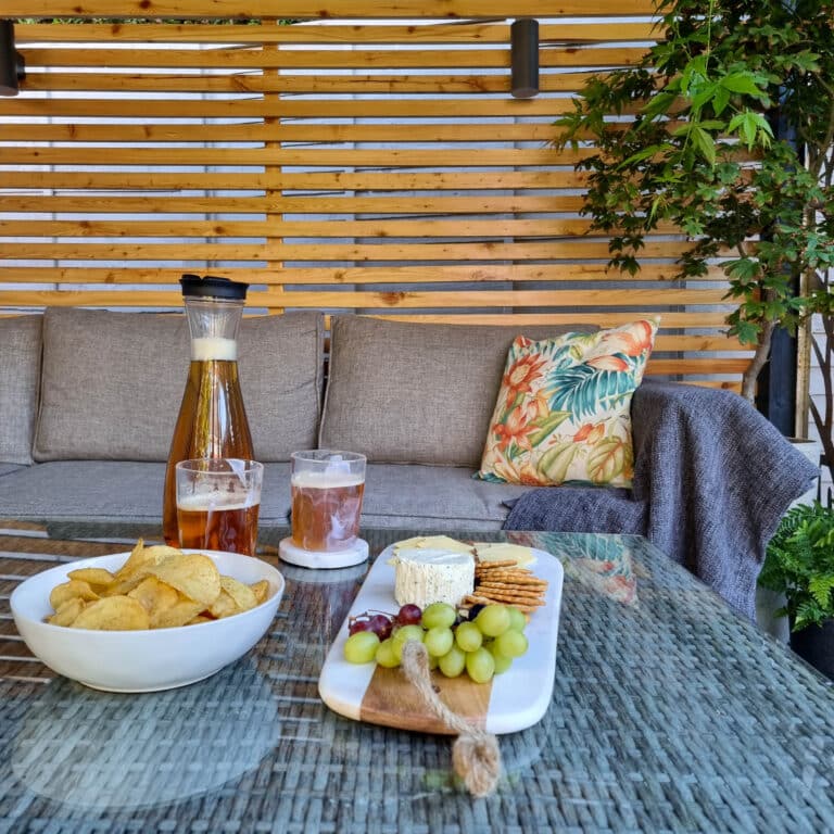 A cosy outdoor patio by a local gate company features a grey sofa, tropical cushion, and throw blanket. On the glass table are a cheese board, crisps, iced tea, and a carafe. Lush green plants add a fresh touch to this inviting space.