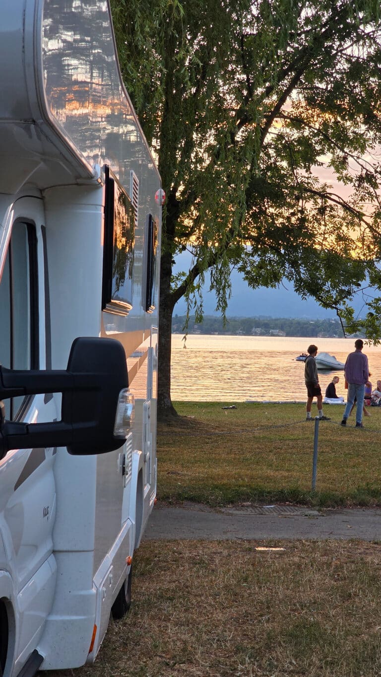 A white camper van is parked near a grassy lakeside at sunset. In the background, a family enjoys their first holiday with a baby, relaxing by the water under the shade of a tree, with boats gently floating on the lake.
