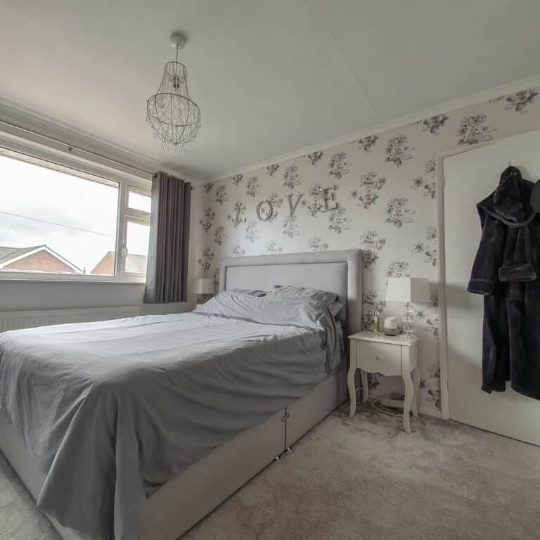 A neatly made bed with grey bedding from Terry’s Fabrics is against a floral wallpapered wall with the word LOVE above the headboard. A white bedside table, lamp, and a dark dressing gown hanging on a door complete the cosy, bright bedroom.