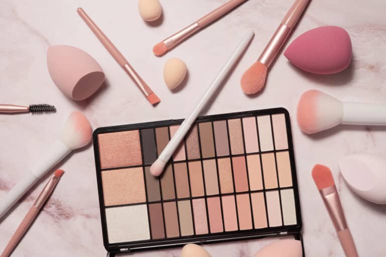 An open eyeshadow palette with neutral shades is surrounded by vegan make-up brushes, blending sponges, and applicators on a light pink marble surface.