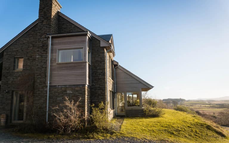 Modern stone and wood house on a grassy hillside with large windows, garden pods nearby, and a clear blue sky, overlooking a scenic rural landscape and distant hills on a sunny day.