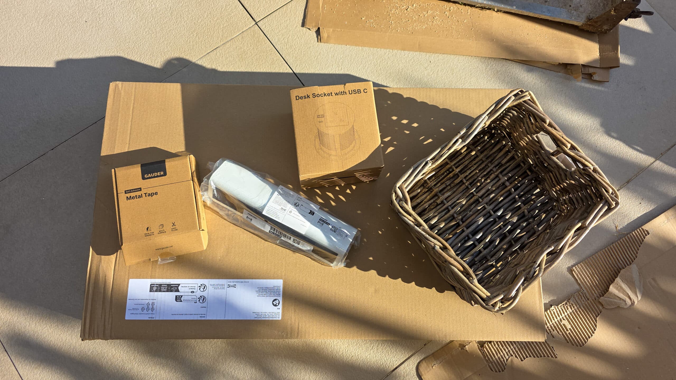 IKEA Malm Bedside Table Hack With Phone Charging. A cardboard sheet with a wicker basket, a box labelled Desk Socket with USB C, a box labelled Metal Tape, an electrical power strip in plastic, and an instruction sheet, all arranged in sunlight.