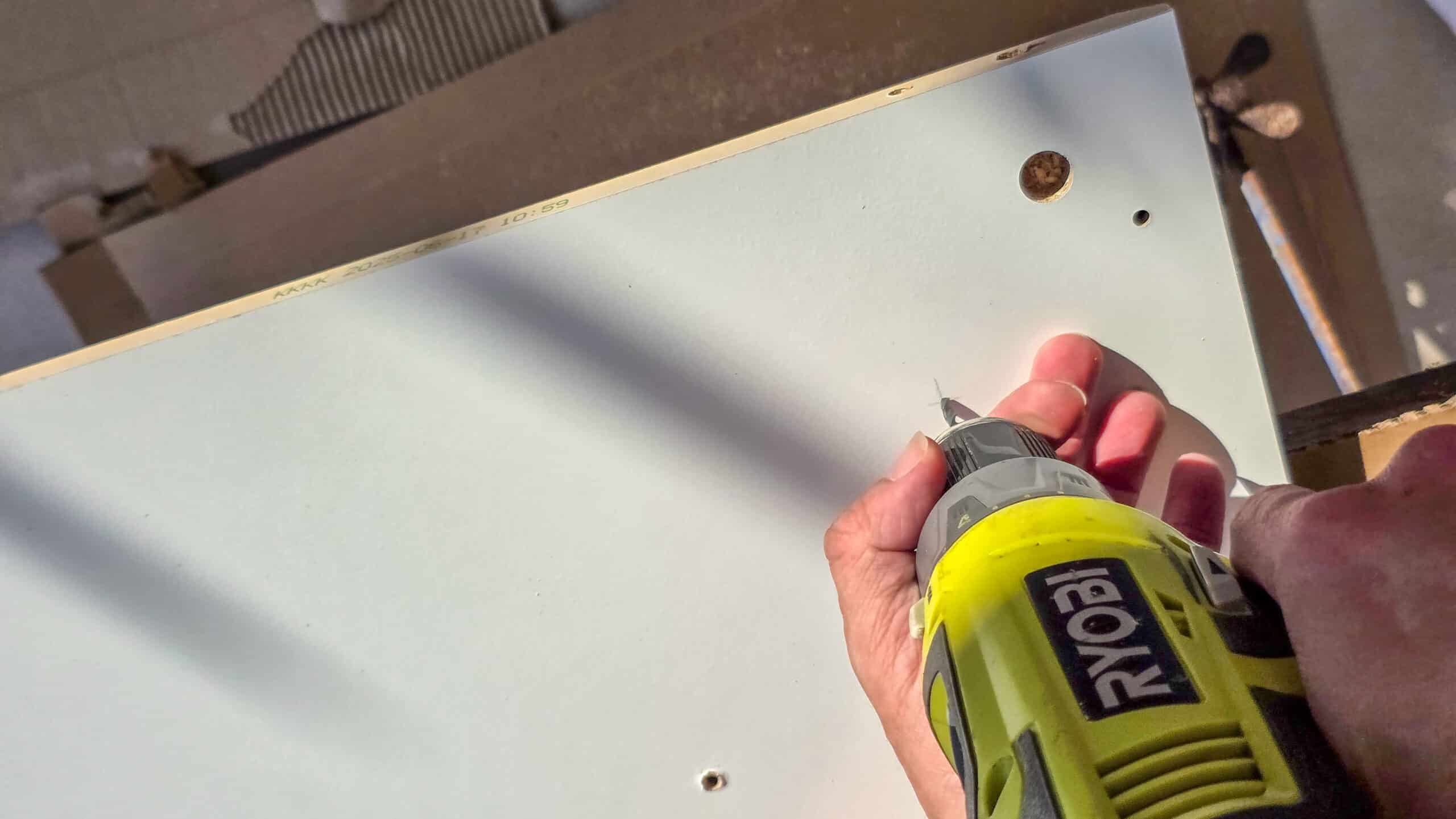 A person uses a Ryobi power drill to drive a screw into a white wooden board, with sunlight casting diagonal shadows across the surface.