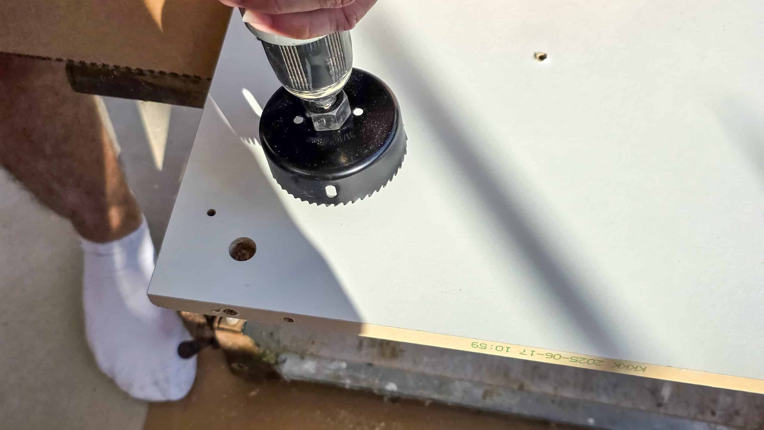 A person uses a hole saw drill bit to cut a circular hole in a white wooden board. Nearby holes and measurement markings are visible, and the person is wearing white socks.