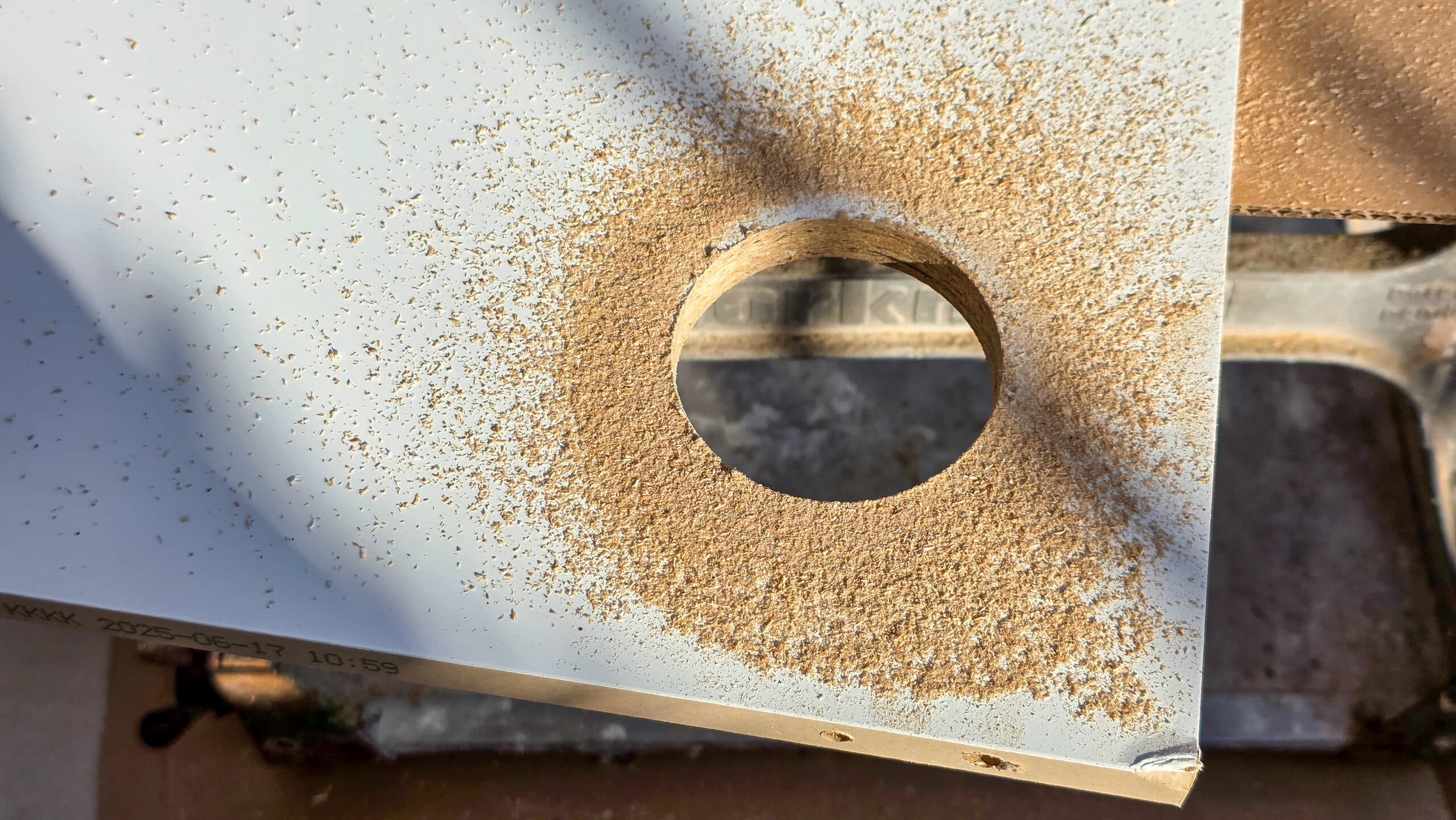 A close-up of a white board with a large circular hole drilled through it, surrounded by scattered sawdust. Sunlight casts shadows across the surface.