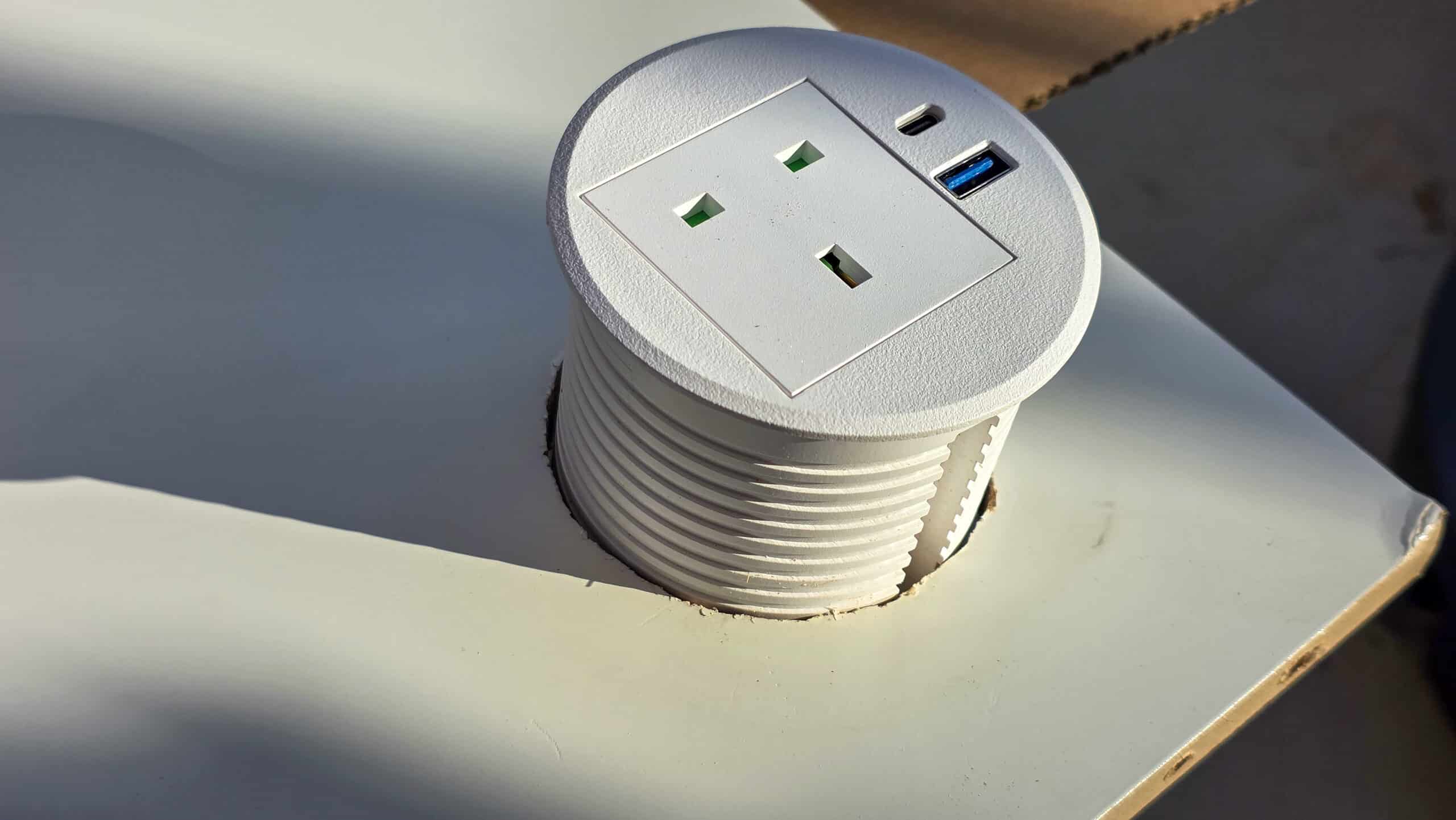 A close-up of a round, white pop-up power outlet installed in a desk, featuring two power sockets and two USB ports. Sunlight casts shadows on the surface around the outlet.