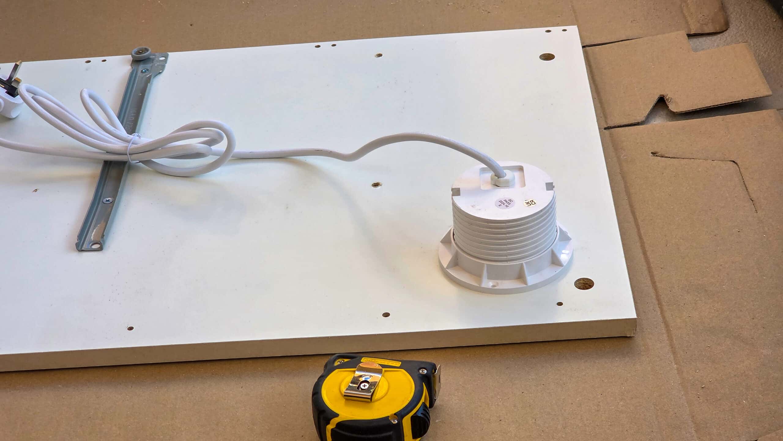 A white electrical fitting with a lead is mounted on a flat white board, with a yellow tape measure and pieces of cardboard visible nearby. The setup suggests ongoing installation or assembly work.