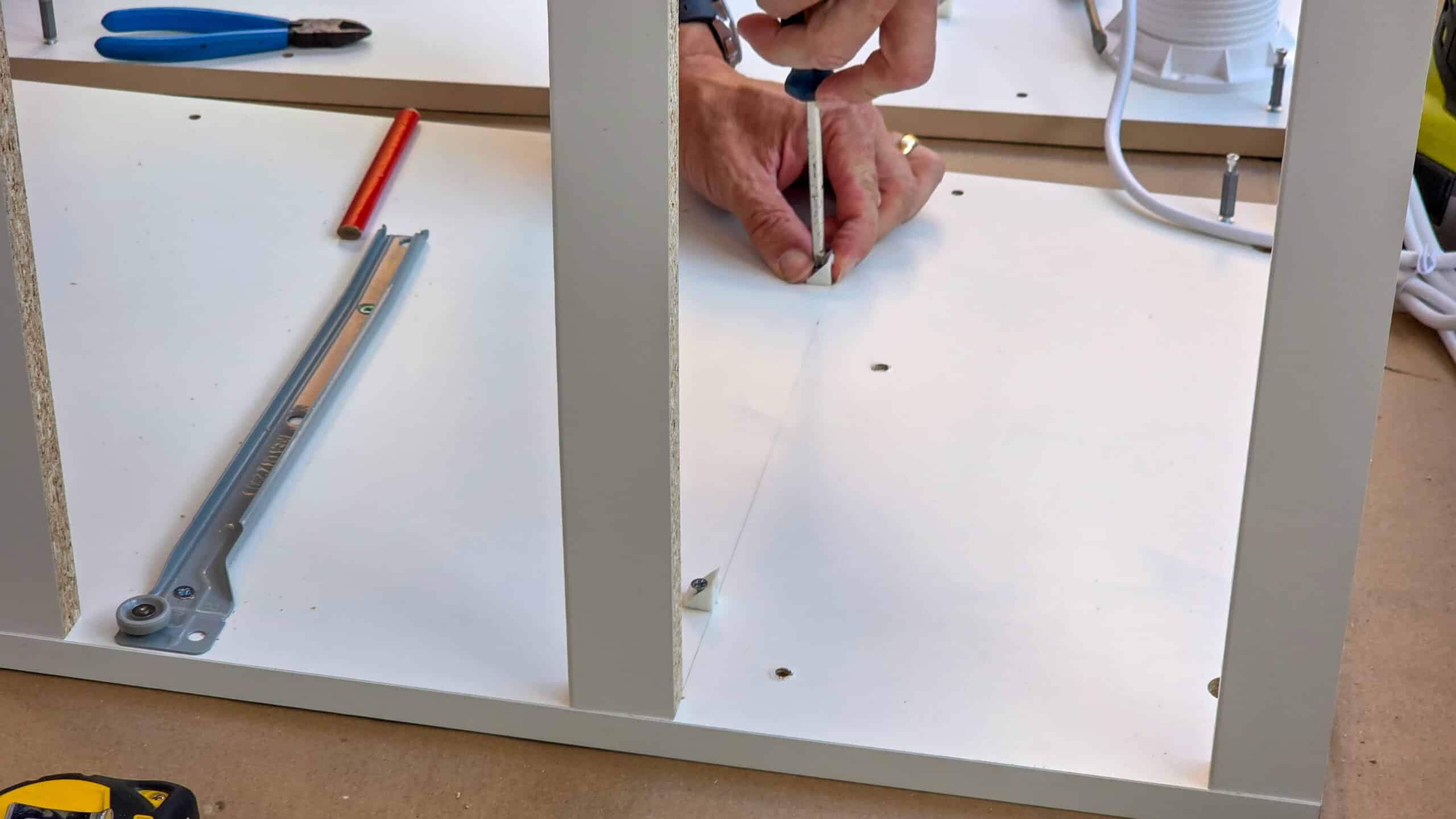Close-up of hands using a screwdriver to assemble white flat-pack furniture, with tools and hardware like a pencil, ruler, and drawer runner visible on the surface.